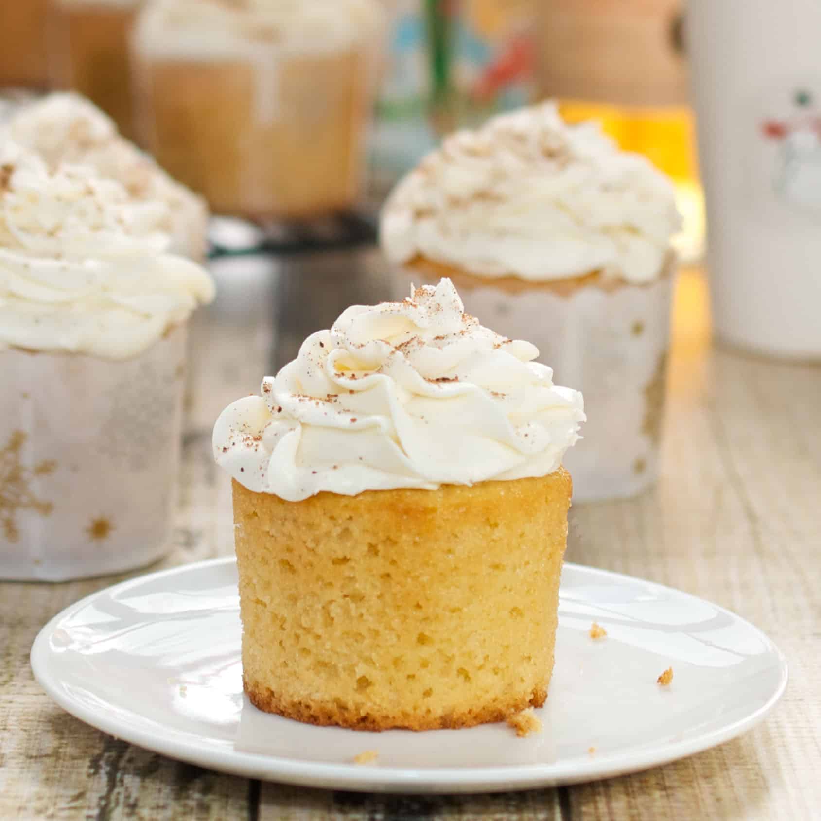 An unwrapped eggnog cupcake on a white saucer with more cupcakes in the background